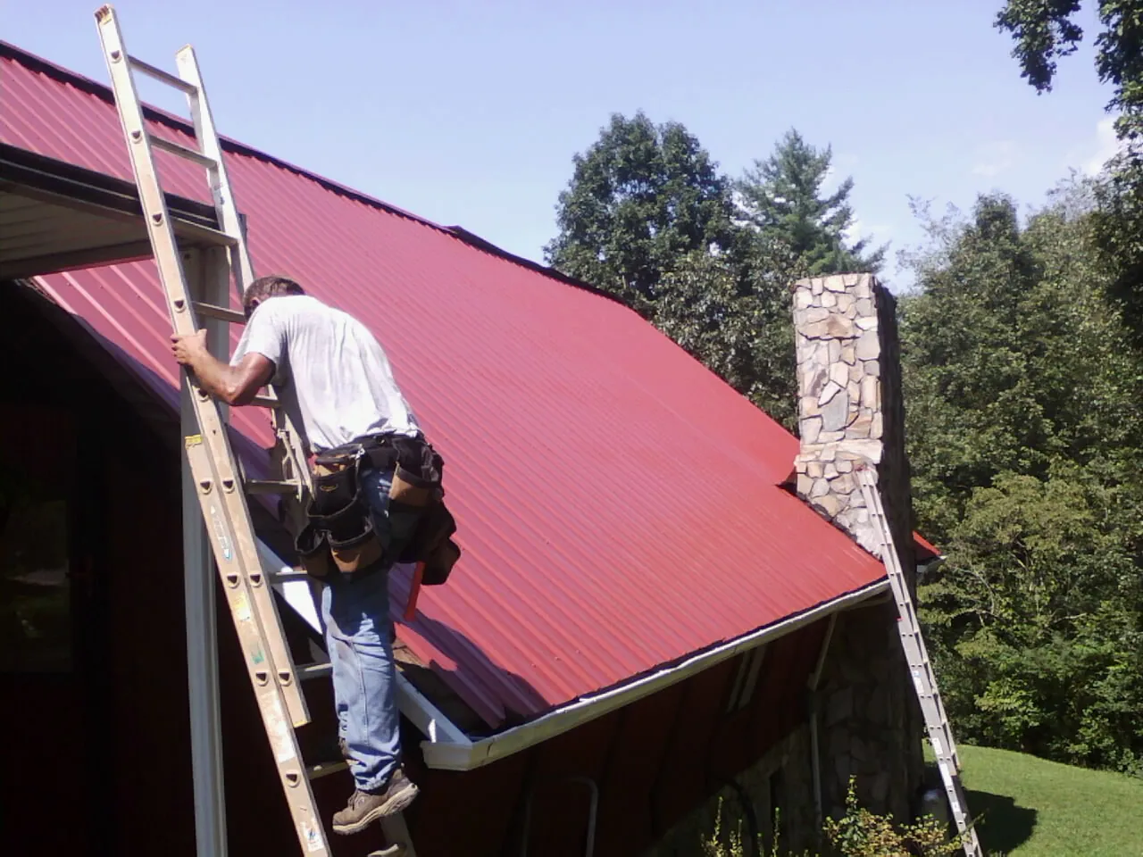 Roofer on a ladder beside a red metal roof and stone chimney — Soffit & Fascia Repair by Roofing Brothers in Laurier